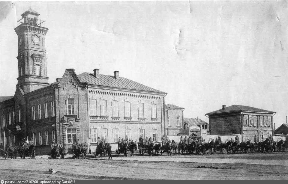 Чёрно-белая фотография здания пожарной части с каланчой и конными пожарными расчётами на площади